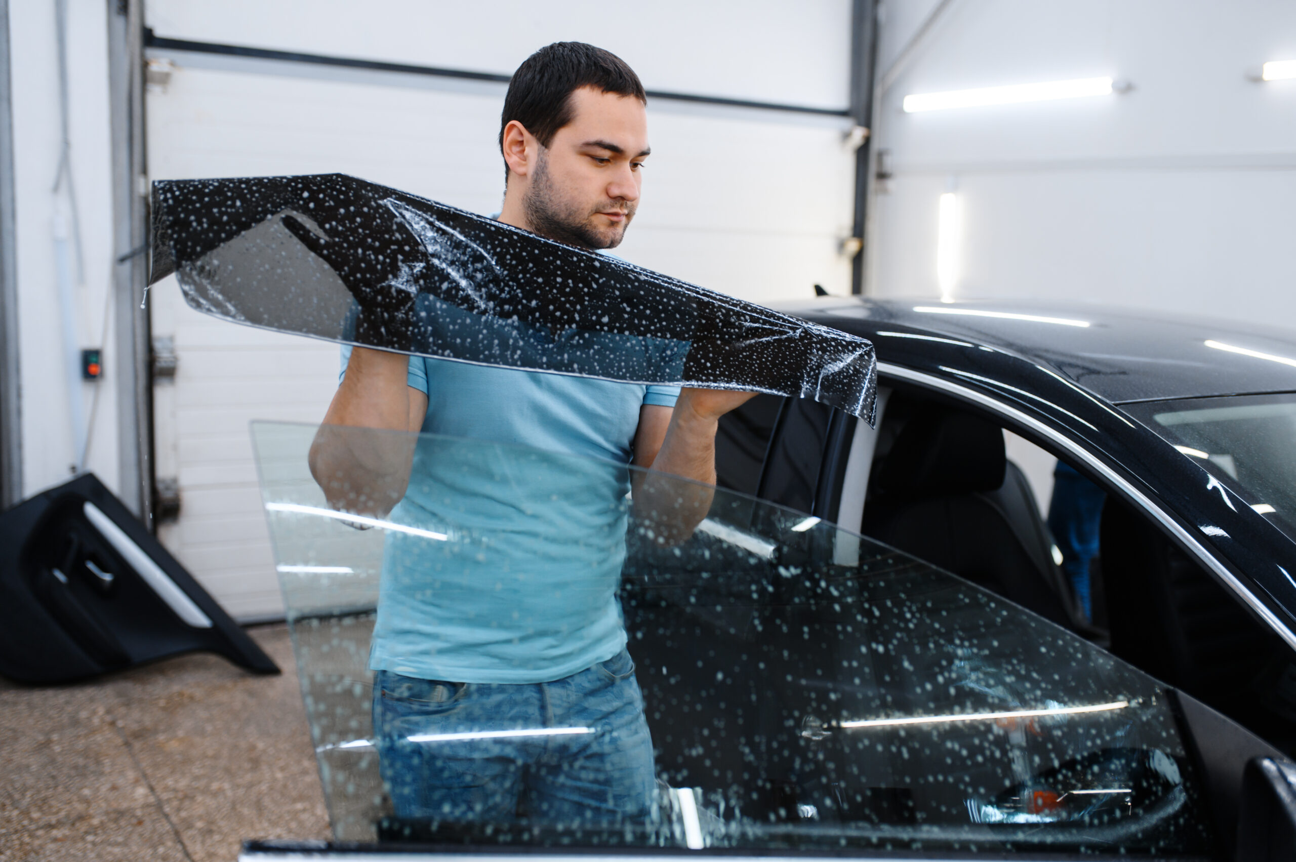 Male specialist installs wetted car tinting, tuning service. Mechanic applying vinyl tint on vehicle window in garage, tinted automobile glass